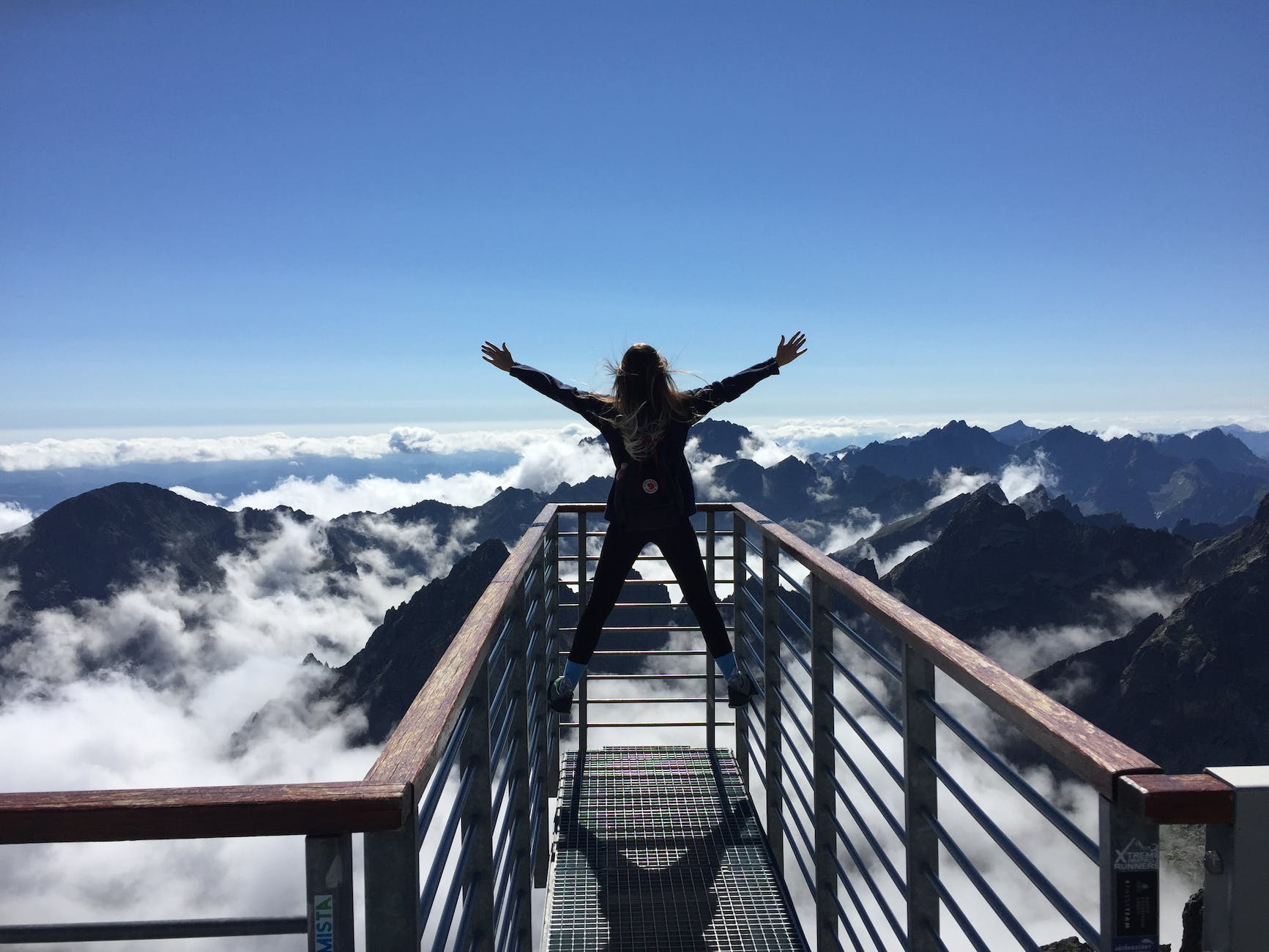 person standing on hand rails with arms wide open facing the mountains and clouds